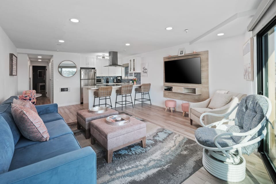 Living room with smart TV, comfortable couch and 2 lounge chairs. Kitchen with bar seating, 2-person dining table, and electric stove/range.