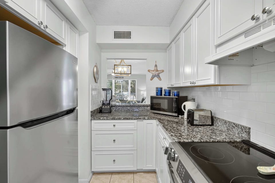 The kitchen is a bright and stylish space, perfect for preparing delicious meals. It features granite countertops, a coffee maker, a refrigerator, an oven, a stove, a microwave, a dishwasher, a toaster, a kettle, cookware, dishes and silverware. 