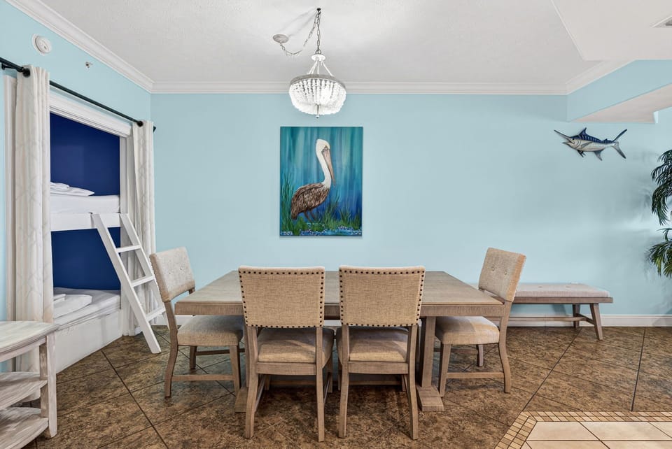 Dining area with a wooden table, chairs, and a bunk bed in a light blue room featuring coastal artwork.