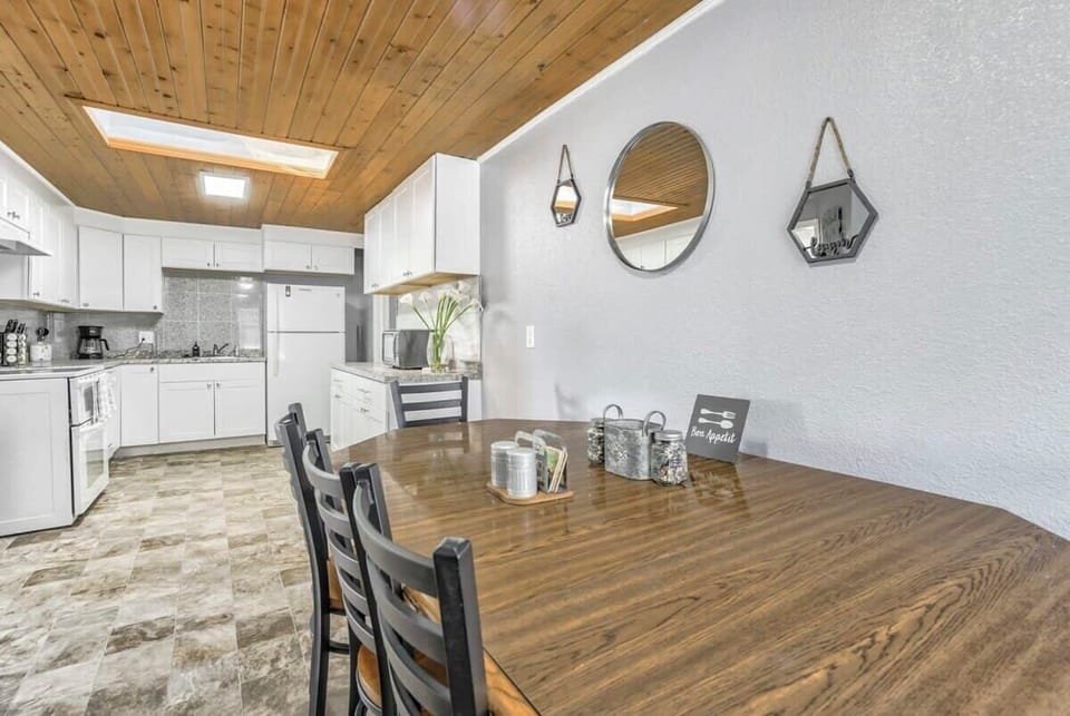 A wood plank ceiling draws the eye above this dining area where a long table invites shared meals and conversation. The open view into the white kitchen keeps everything connected while warm tones create a welcoming place to gather.