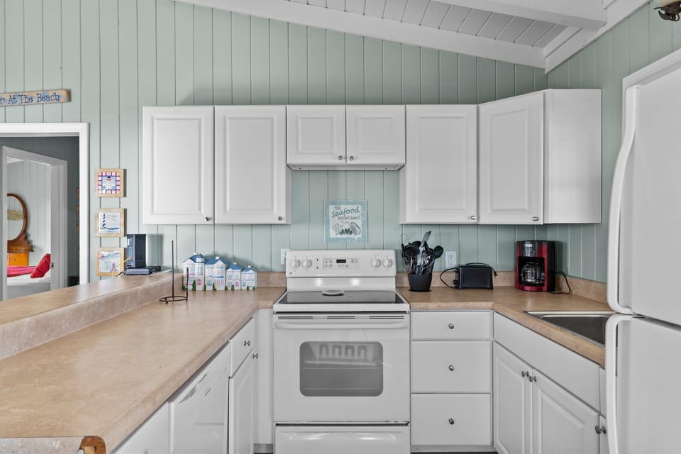 Open kitchen with white cabinets, full fridge, stove, sink, coffee maker, breakfast bar stools, vaulted ceiling, beach décor, and airy Oak Island, NC style.