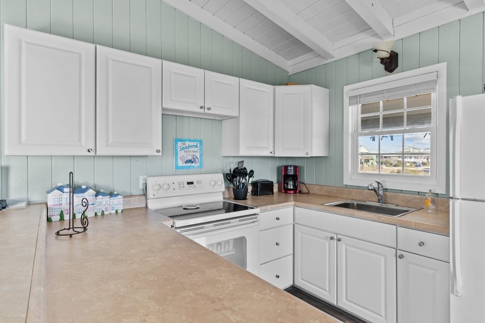 Open kitchen with white cabinets, full fridge, stove, sink, coffee maker, breakfast bar stools, vaulted ceiling, beach décor, and airy Oak Island, NC style.