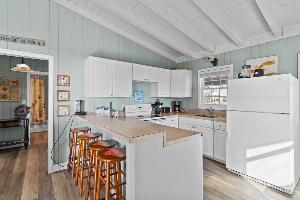 Open kitchen with white cabinets, full fridge, stove, sink, coffee maker, breakfast bar stools, vaulted ceiling, beach décor, and airy Oak Island, NC style.