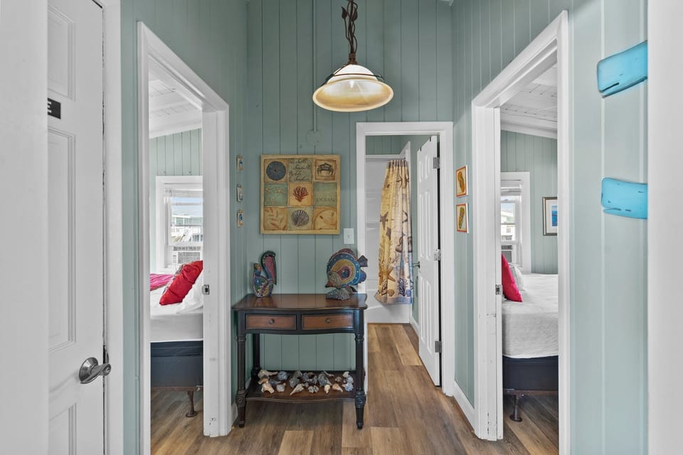 Beachy hallway with seafoam walls, shell art, fish décor, console table, wood floors, pendant light, open bedrooms, and bath entry in Oak Island, NC.