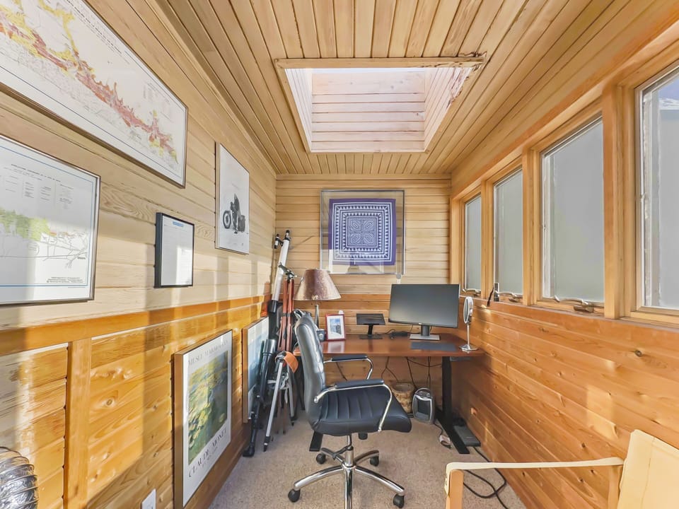 A small office with wooden walls, a skylight, multiple framed pictures, a desk with a computer, a black office chair, and a lamp. There is a window on the right side.