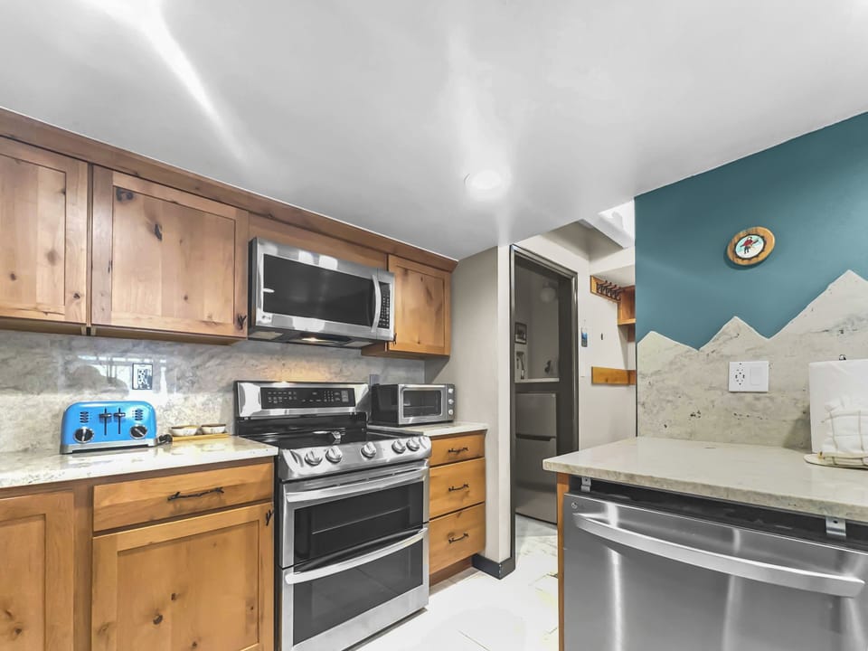 A modern kitchen featuring wooden cabinets, a stainless steel stove, microwave, dishwasher, a blue toaster, and a marble countertop with a mountain-themed wall design in the background.