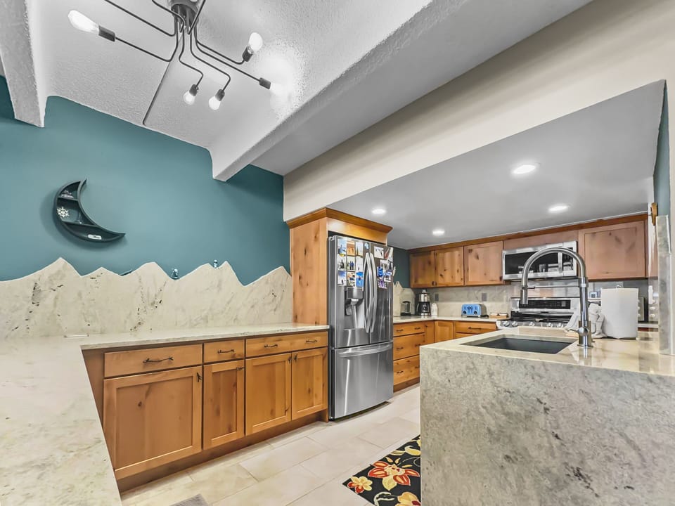 Modern kitchen with wooden cabinets, a stainless steel refrigerator, and a marble countertop. A decorative moon clock is on a teal accent wall, and the ceiling lights are contemporary.