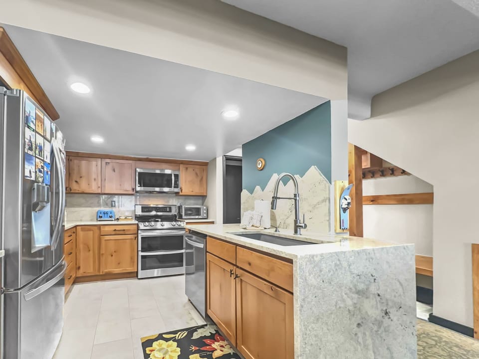 Modern kitchen with wooden cabinets, stainless steel appliances, and a large island with a sink. Refrigerator has magnets and photos. Stairs are visible on the right side.