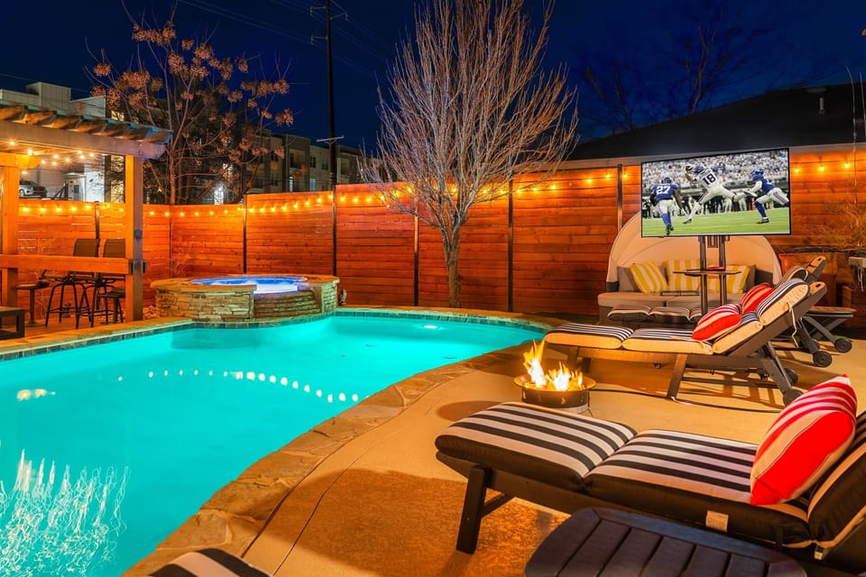 Nighttime view of a backyard with a glowing pool and hot tub, lounge chairs facing a football game on an outdoor TV, a fire pit, string lights, and a covered patio area.