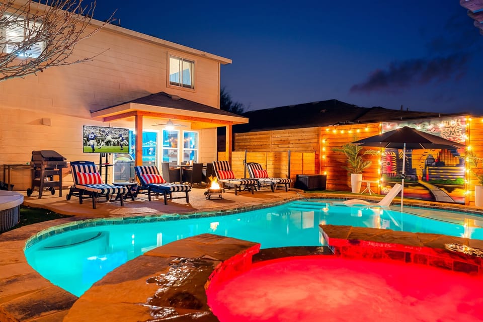 Nighttime view of a backyard oasis featuring a glowing pool and hot tub, lounge chairs facing a TV, a grill, string lights, and a mural on the fence.