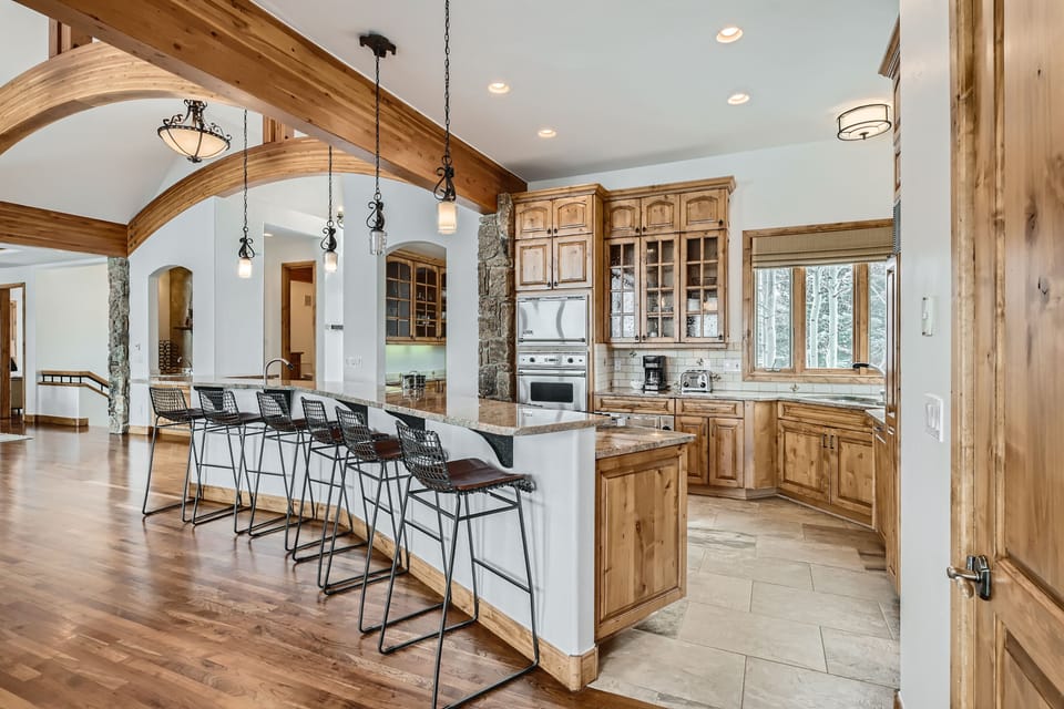 Modern kitchen with wooden cabinets, a curved granite countertop island, and four black barstools. The space features pendant lighting, stainless steel appliances, and a mix of wood and tile flooring.