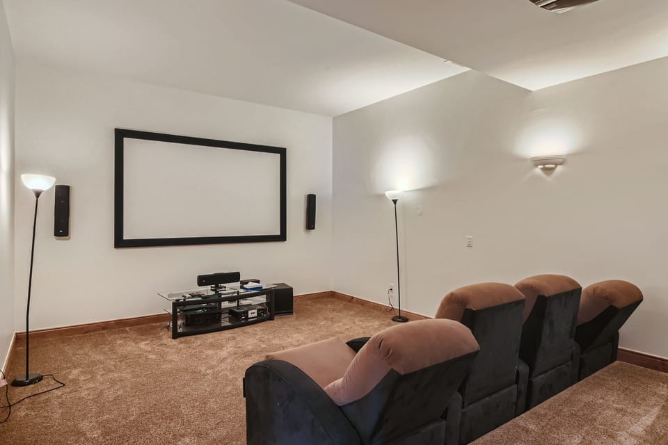A home theater room with three cushioned seats, a large projector screen on the wall, two floor lamps, and a media stand with electronic devices underneath the screen. The room is carpeted.