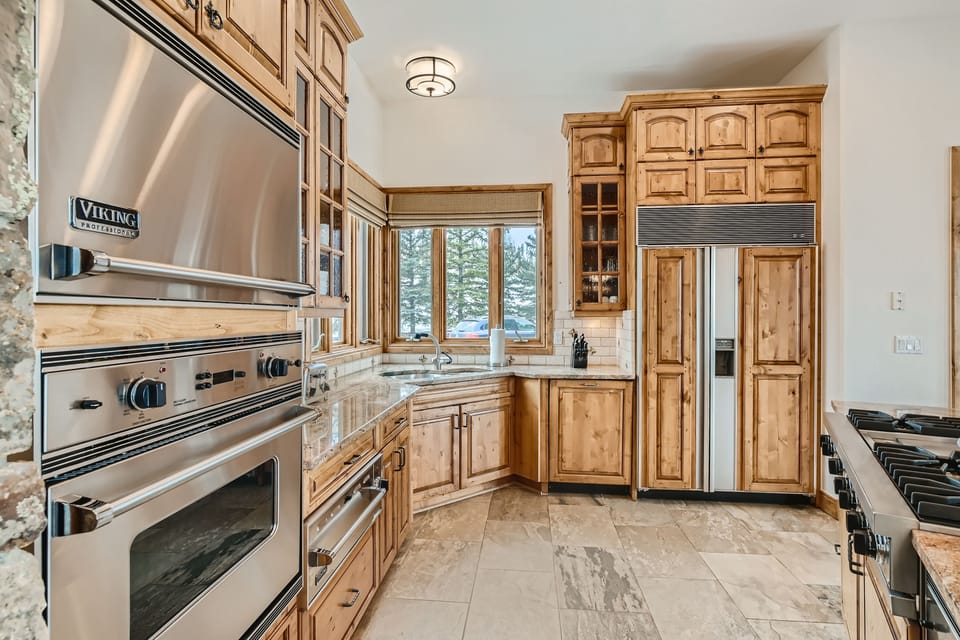 A modern kitchen with stainless steel appliances, wooden cabinets, a double-door refrigerator, an oven, a stove, and a large window overlooking a snowy landscape.