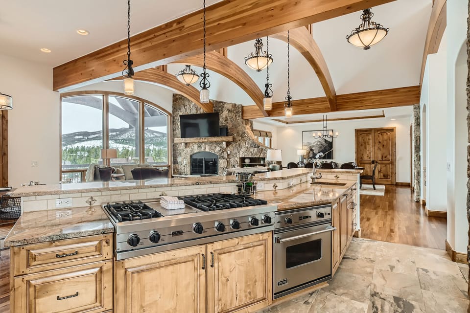 A spacious kitchen with wooden beams and granite countertops, featuring a gas stove, double ovens, and a stone fireplace in the background. Large windows overlook a scenic outdoor view.