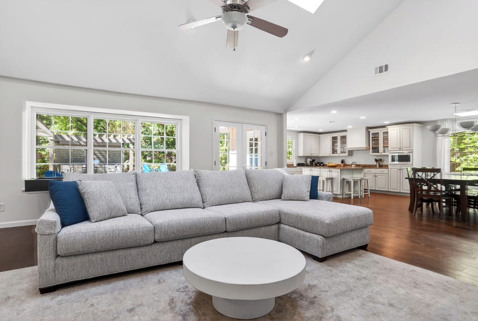 Open‑concept living space with soft neutrals, vaulted ceilings, and sunlit views flowing seamlessly from living room to kitchen and dining.