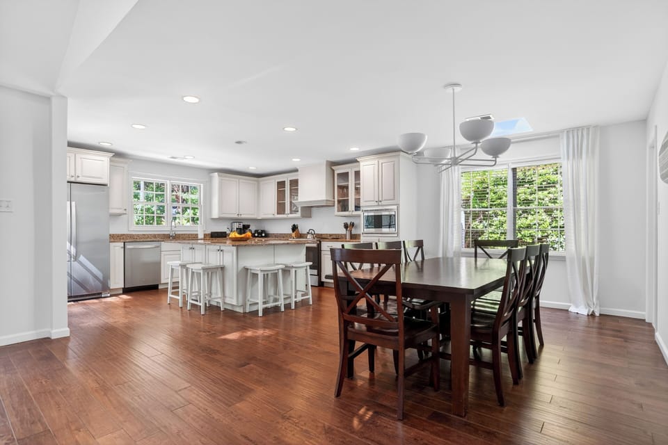 Bright, modern kitchen and dining space with white cabinetry, granite counters, and sun‑filled windows overlooking greenery.