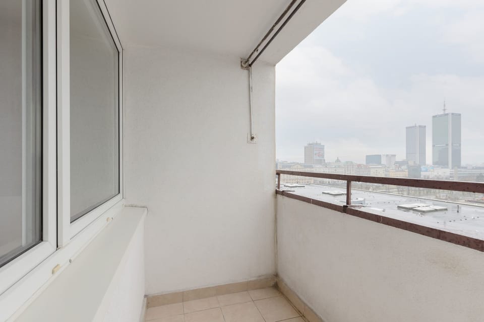 A balcony view overlooking a cityscape. The balcony has a clean, minimalist design with a metal railing and tiles on the floor, and it offers a distant view of the city.