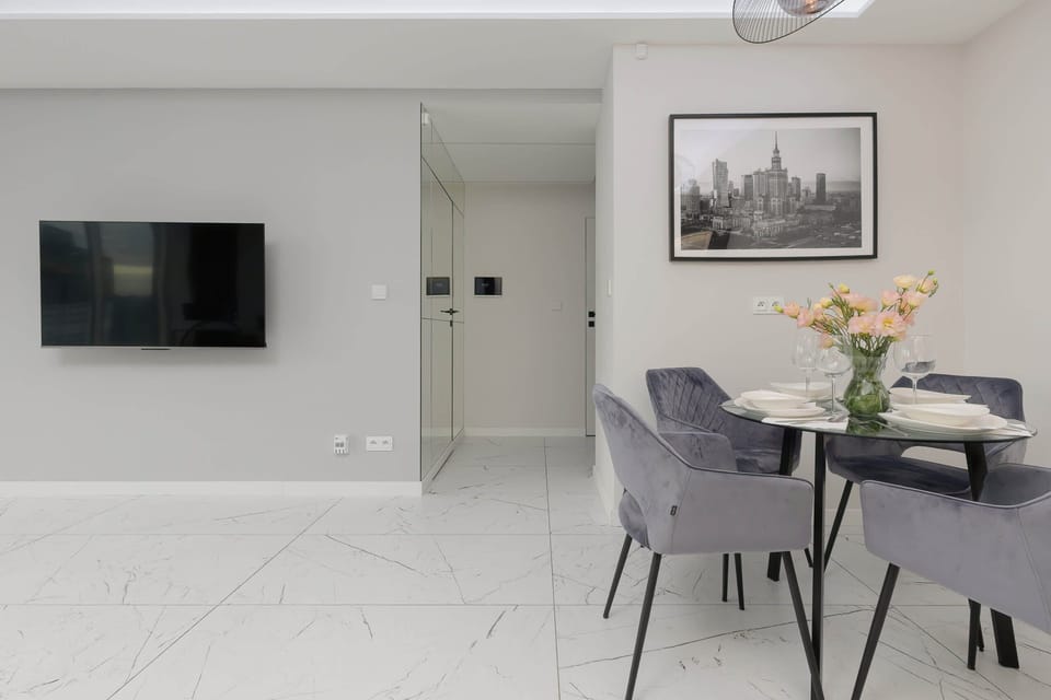 An elegant living area featuring a black leather sofa set, a marble coffee table, and a flat-screen TV mounted on the wall, with soft natural light streaming in through large windows.