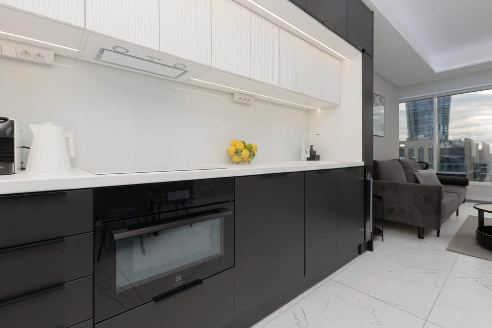 A minimalist kitchen with white cabinets and black countertops, featuring modern appliances, and a large window that allows natural light to brighten the space.