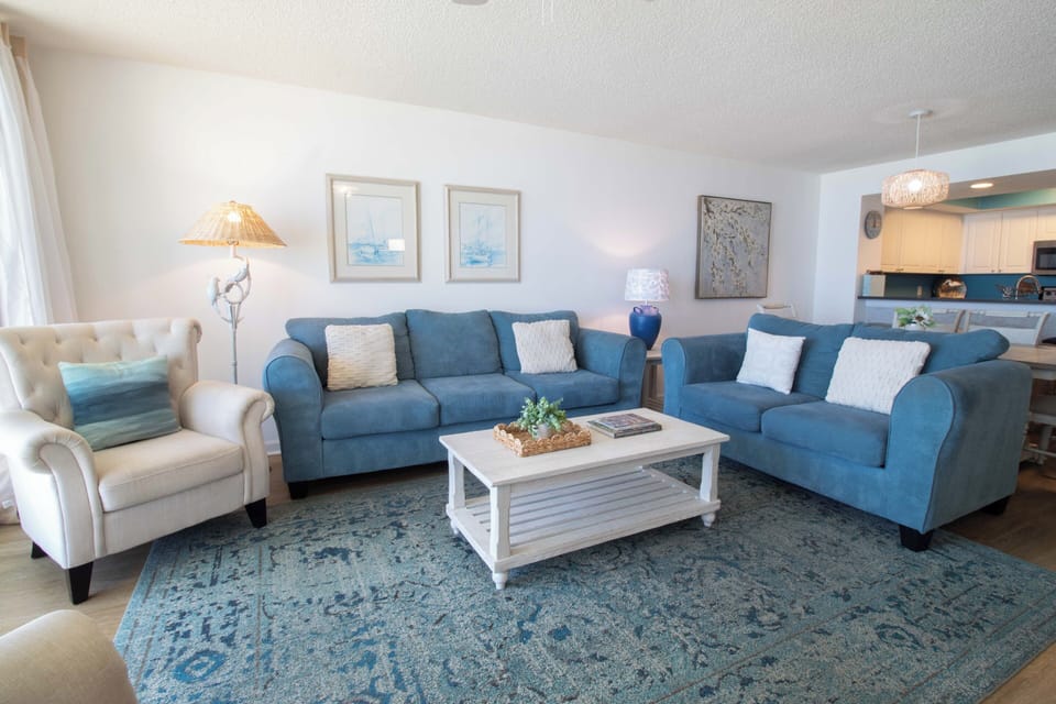 Relax in your spacious living room with plush blue sofas, coastal artwork, and open kitchen views—perfect for unwinding after beach days.