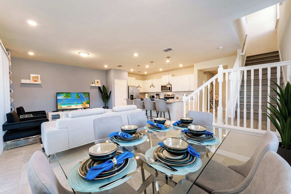 Open-plan dining area with a glass table set for six, blue napkins, and a bright white kitchen with bar seating.