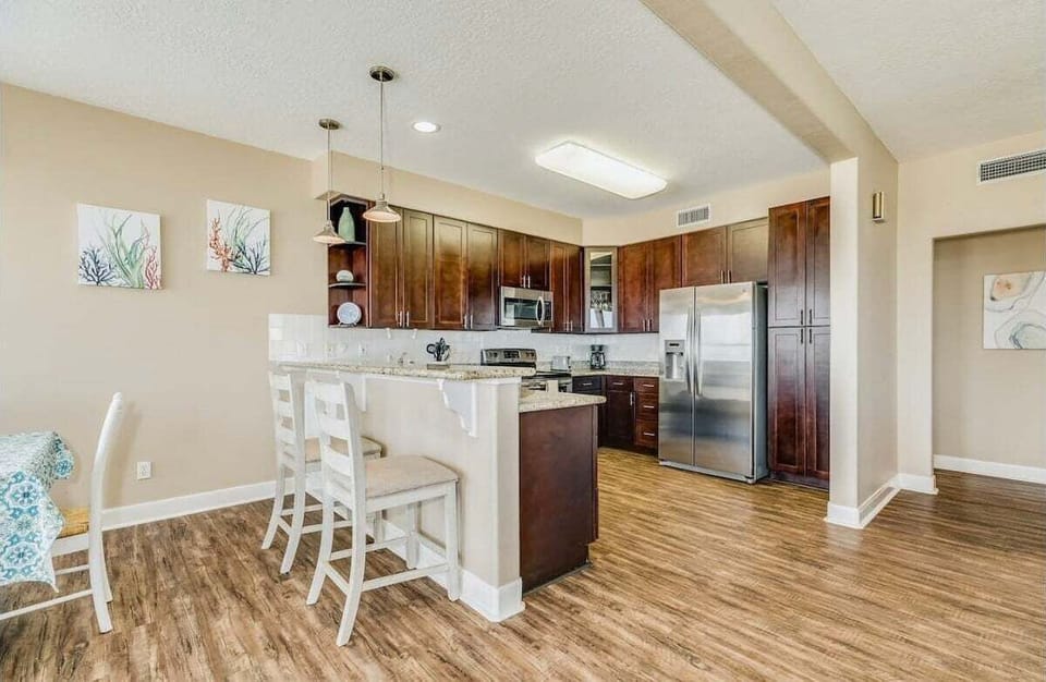 Bright kitchen and dining area with large island, bar stools, stainless appliances, wood cabinetry, and open layout.