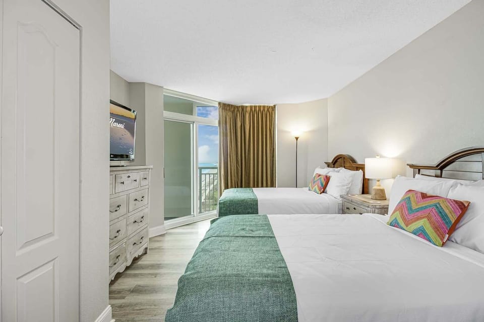 Comfortable bedroom with two beds, colorful coastal accents, TV, dresser storage, and balcony access with a peek of the ocean.
