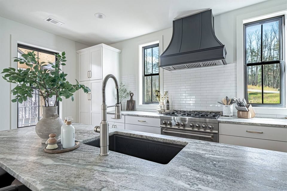 Bright, modern kitchen with a large granite island, deep sink, and sleek gooseneck faucet. White cabinetry, a black statement range hood, and professional style stainless gas range are framed by tall windows that fill the space with natural light.