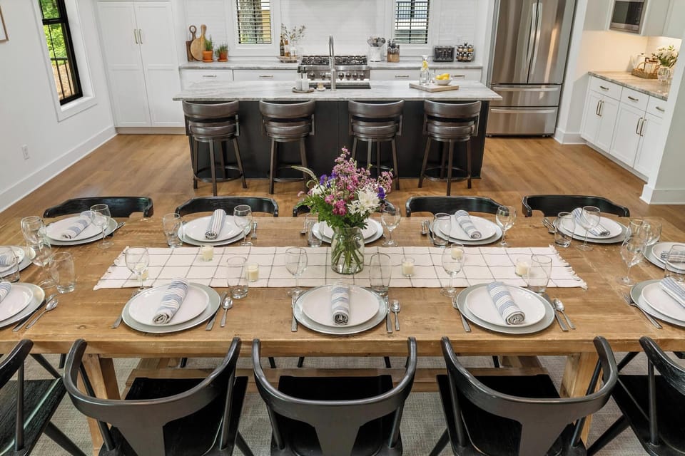Spacious dining area with a reclaimed wood table that seats 12. Behind it, a large kitchen island with 4 bar stools, stainless appliances, and bright cabinetry creates an open, social space for gathering.