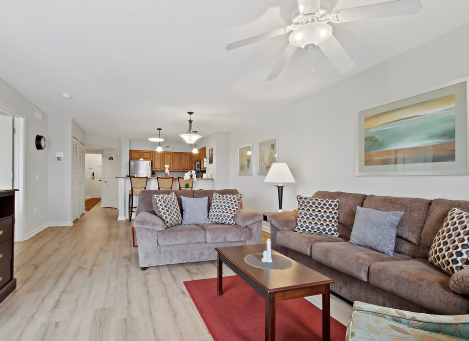 Settle into your spacious living room where plush sofas invite relaxation under gentle ceiling fan breezes, with open sightlines to your fully equipped kitchen.