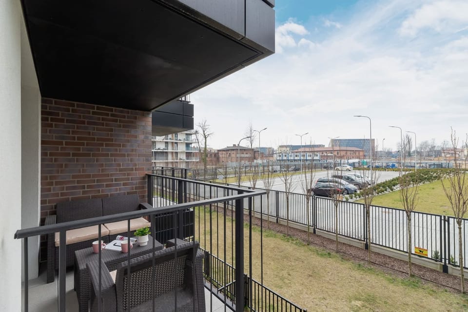 A balcony with a black railing, offering a view of a green space and modern buildings in the distance. A relaxing spot for fresh air.