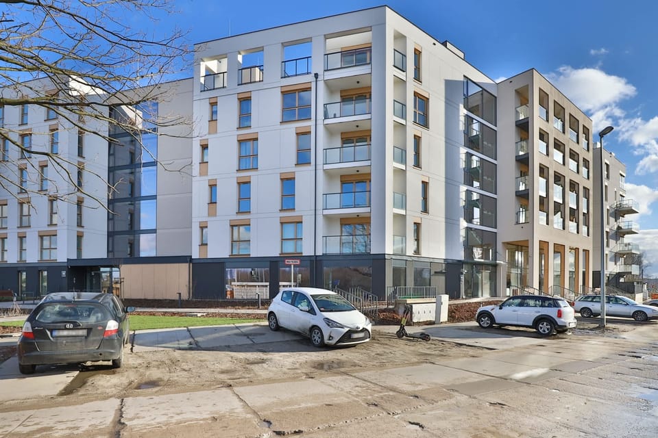 An exterior view of the modern apartment building under clear blue skies. A clean, well-maintained facade with accessible parking is visible.