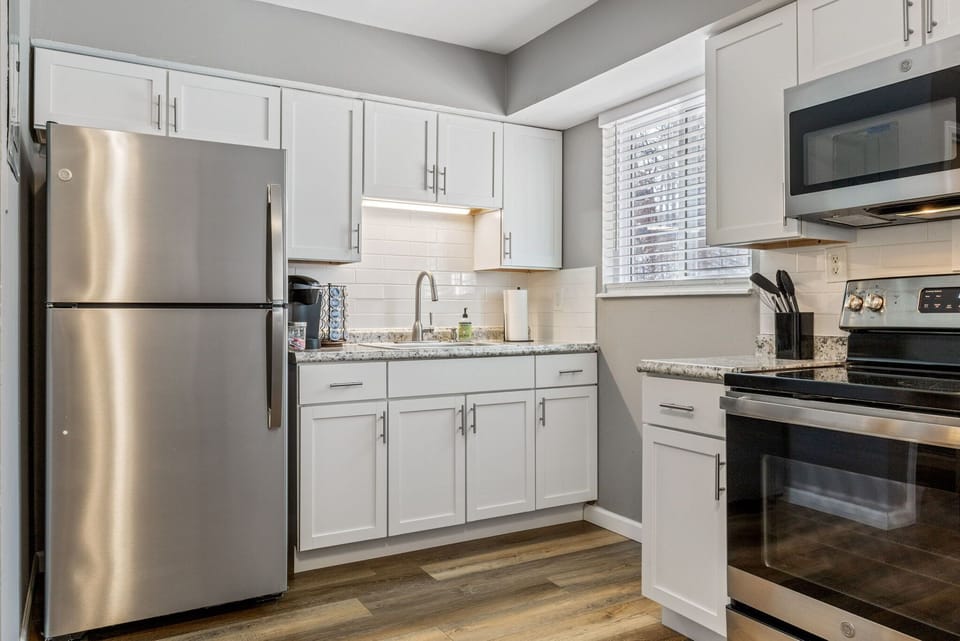 This kitchen view shows the stainless-steel refrigerator, range, and microwave alongside white cabinetry, counters, and a window.