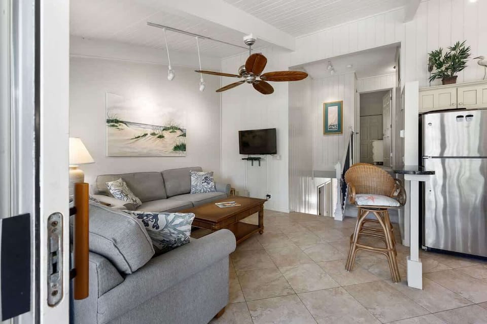 Sink into the cozy sectional and enjoy your coastal retreat with ocean-themed art, tropical ceiling fan, and modern kitchen nearby.