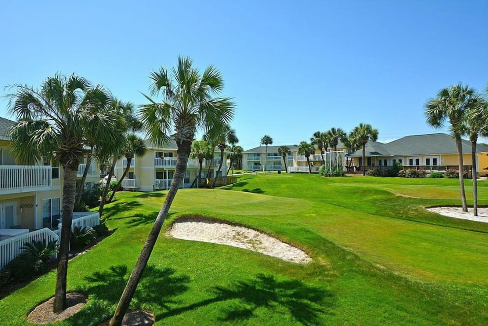 Golf course with sand bunkers and palm trees, surrounded by resort buildings under clear blue skies.