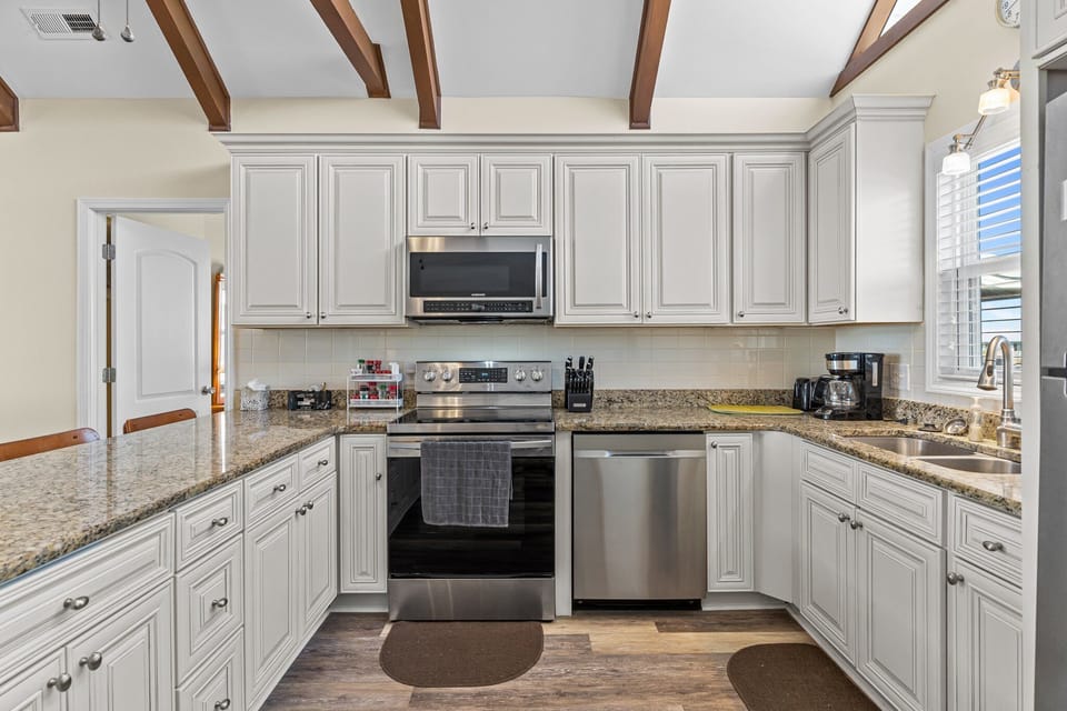 Open kitchen with granite island seating for four, white cabinets, stainless appliances, vaulted ceiling with exposed beams, ceiling fan, and large windows bringing in natural light.