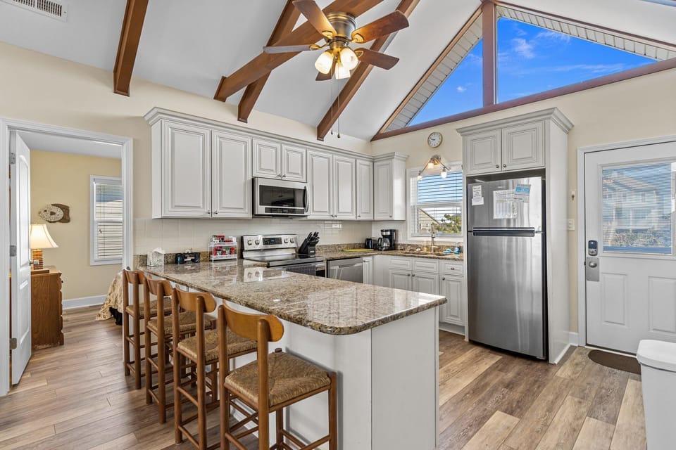 Open kitchen with granite island seating for four, white cabinets, stainless appliances, vaulted ceiling with exposed beams, ceiling fan, and large windows bringing in natural light.