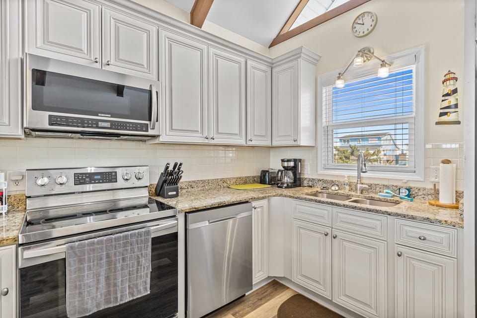 Open kitchen with granite island seating for four, white cabinets, stainless appliances, vaulted ceiling with exposed beams, ceiling fan, and large windows bringing in natural light.