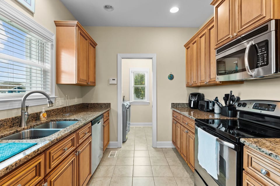 Kitchen with granite countertops, stainless steel appliances, double sink, wood cabinetry, tile flooring, and dining area. Bright, fully equipped gathering space in a comfortable Oak Island, NC beach home.