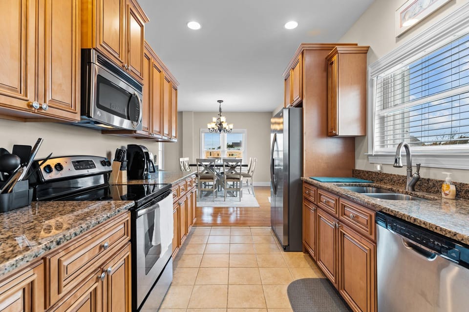 Kitchen with granite countertops, stainless steel appliances, double sink, wood cabinetry, tile flooring, and dining area. Bright, fully equipped gathering space in a comfortable Oak Island, NC beach home.