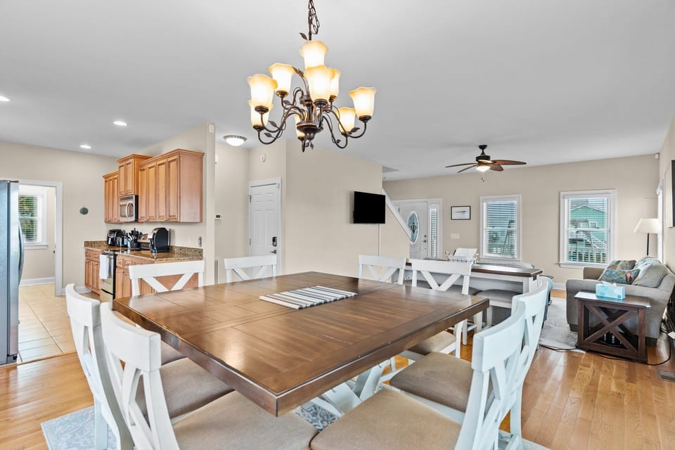 Dining area with table seating for six, chandelier lighting, hardwood floors, large windows, and glass doors leading to the deck. Bright gathering space with coastal views in an Oak Island, NC beach home.