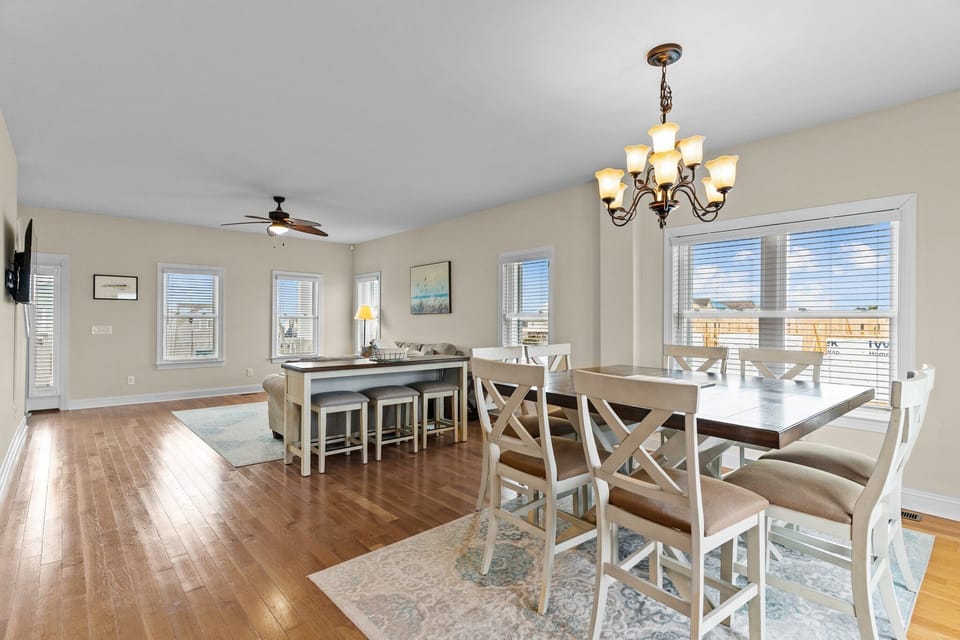 Dining area with table seating for six, chandelier lighting, hardwood floors, large windows, and glass doors leading to the deck. Bright gathering space with coastal views in an Oak Island, NC beach home.
