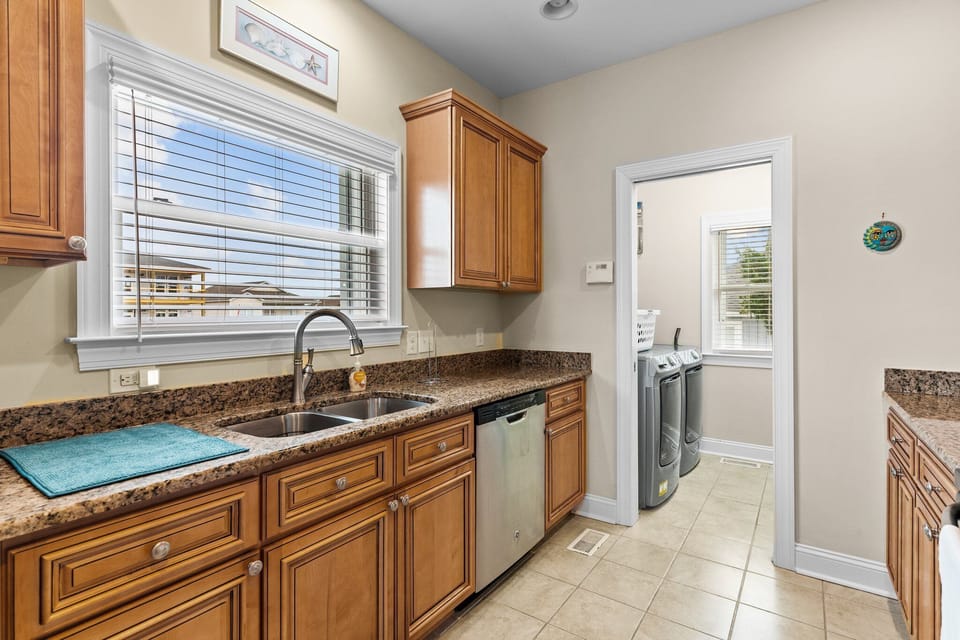 Kitchen with granite countertops, stainless steel appliances, double sink, wood cabinetry, tile flooring, and dining area. Bright, fully equipped gathering space in a comfortable Oak Island, NC beach home.