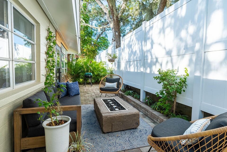 Outdoor patio with brick pavers, outdoor sofa, woven lounge chairs, fire pit coffee table, string lights, tropical plants, and privacy fence