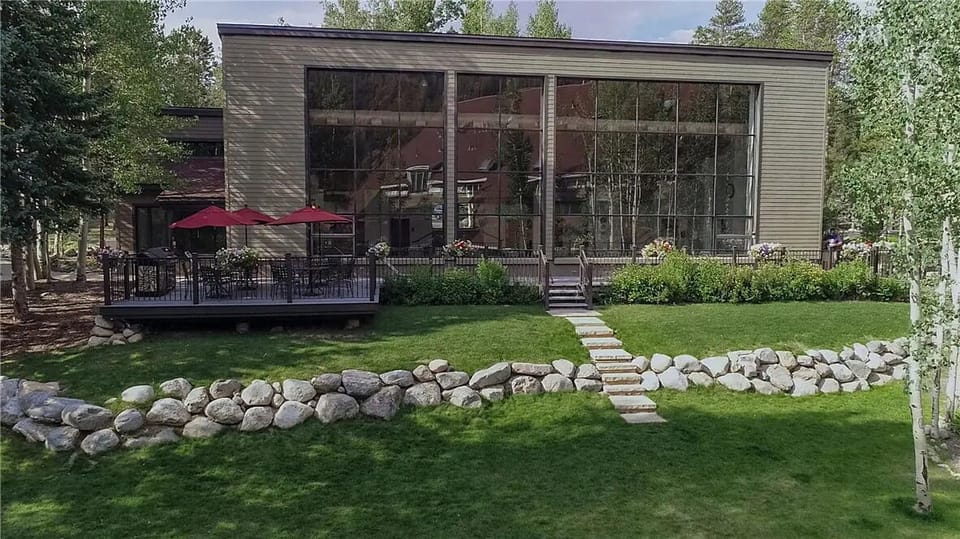 A modern building with large floor-to-ceiling windows, a wooden deck with red umbrellas, and a stone-bordered lawn with a pathway leading to the entrance. Trees surround the structure.