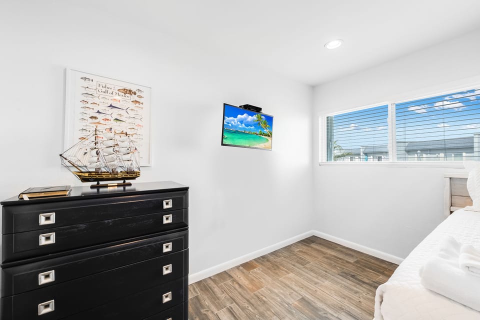 Relax in your bright bedroom featuring a nautical-themed dresser, wall-mounted TV, and natural light streaming through the blinds.
