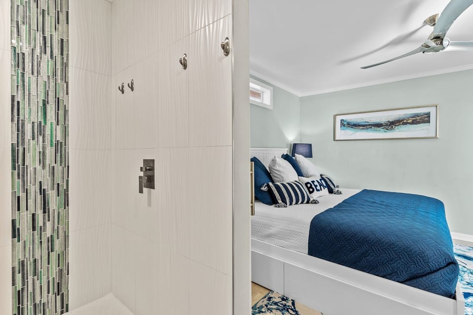 Stylish Oak Island ensuite featuring a spacious walk-in tile shower with vertical glass accent mosaic, modern fixtures, and convenient access directly from the coastal king bedroom with blue bedding and ceiling fan.