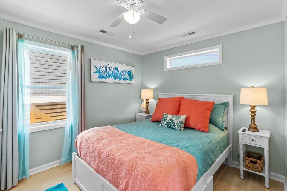Cheerful Oak Island bedroom featuring a white queen bed with coral and teal bedding, sea life artwork, dual nightstands with lamps, ceiling fan, soft green walls, wood-look flooring, and natural light from two windows.