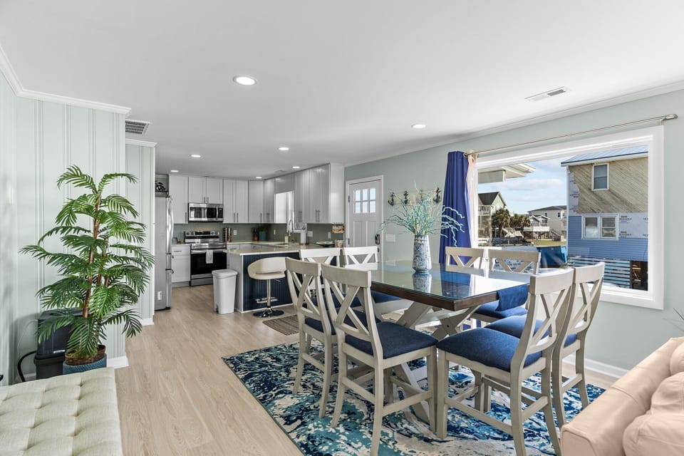 Bright Oak Island open-concept dining and kitchen with seating for six, blue cushioned chairs, coastal rug, stainless appliances, quartz island with bar stools, and large windows filling the space with natural light and beachside views.
