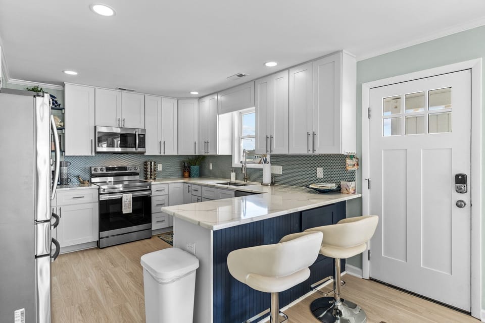 Bright Oak Island kitchen featuring white shaker cabinets, stainless steel appliances, quartz countertops, blue beadboard island with bar seating, mosaic tile backsplash, and natural light from the window and entry door.
